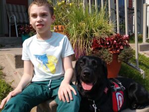 Young boy sits on a stoop with a black lab wearing a red assistance dog cape