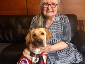 woman sits on a couch with a yellow lab sitting on the floor nxt to her wearing a red cape