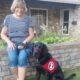 woman sits on a short retaining wall with black lab sitting in the grass wearing a red Can Do Canines service dog cape