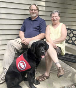 man and woman sitting outside home with black service dog