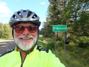 man wearing a bike helmet and bear poses in front of a green road sign that reads the city name of Finland.