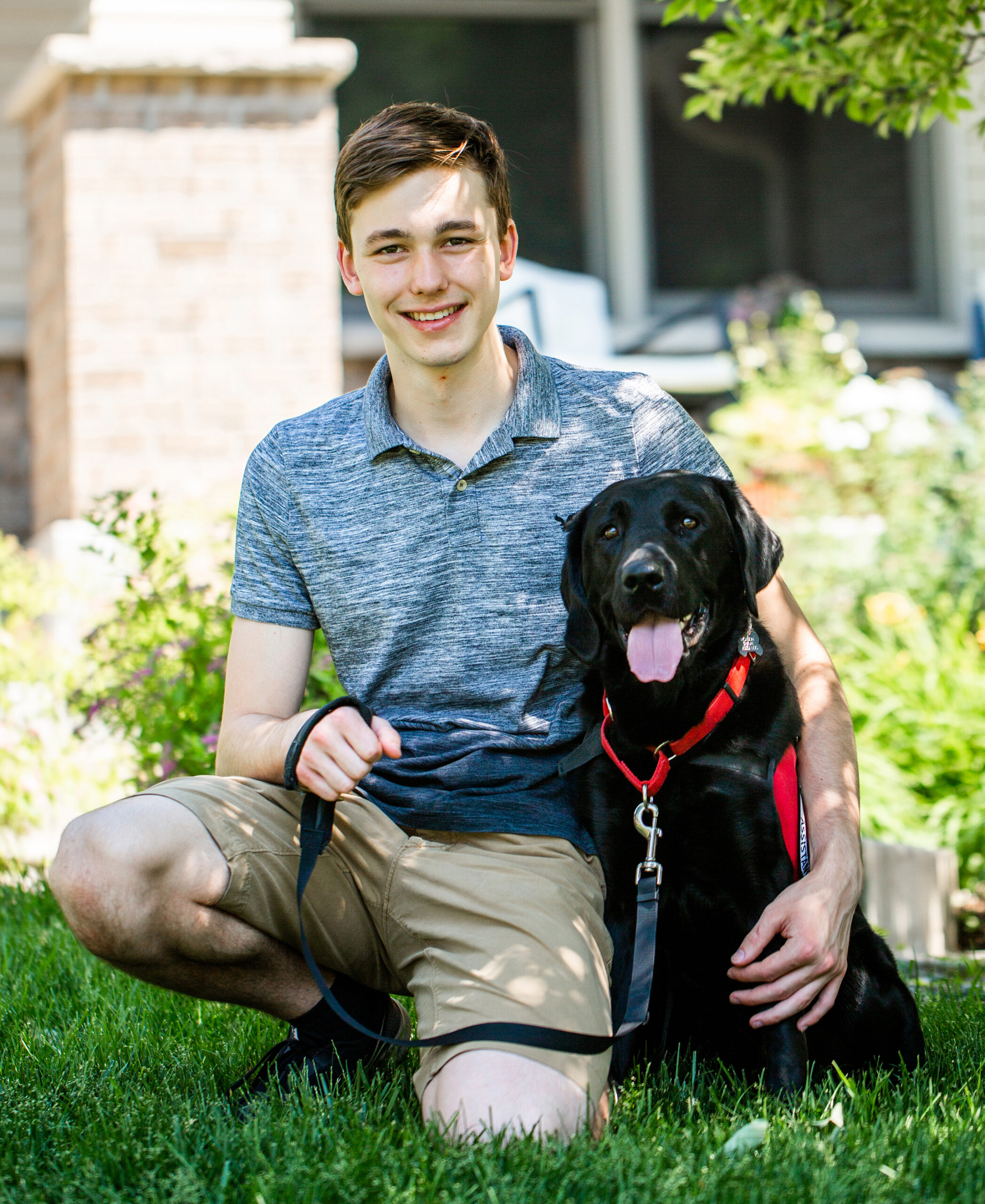 teenage boy kneeling with black service dog wearing a red Can Do Canines cape