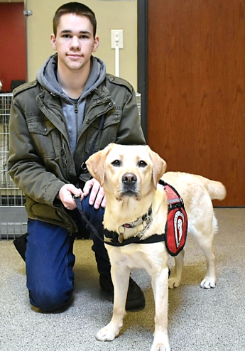 young man kneeling next to yellow Lab service dog wearing a red Can Do Canines cape