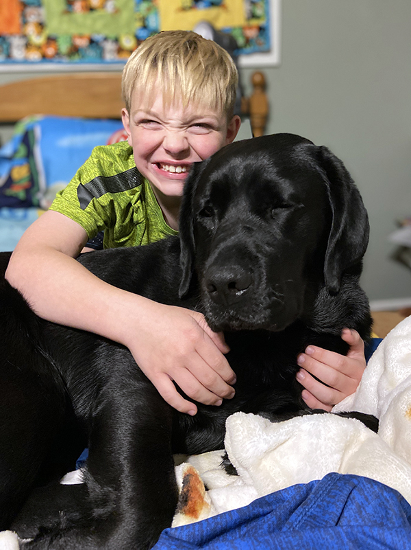 boy with big smile hugging large black dog on bed