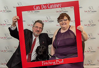 man in suit, woman in dress holding a large frame around them and a black dog