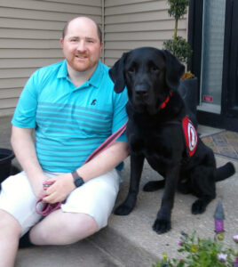 man in turquoise shirt sitting on front steps with black service dog