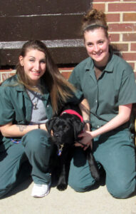 two female prison inmates kneeling outside with black puppy between them