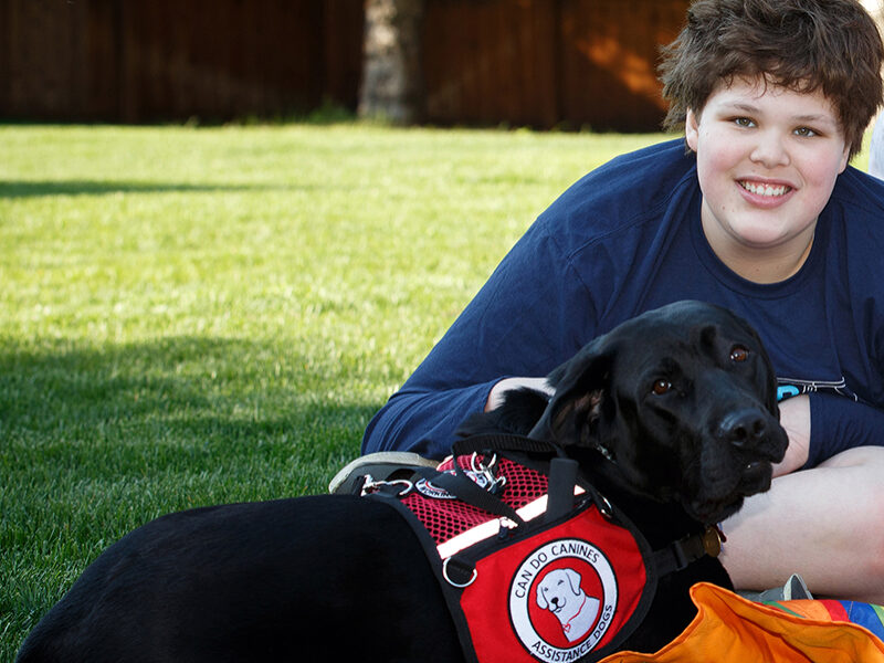 boy sitting on grass with black service dog next to him