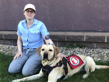 woman kneeling outside with hand on yellow Lab service dog lying next to her