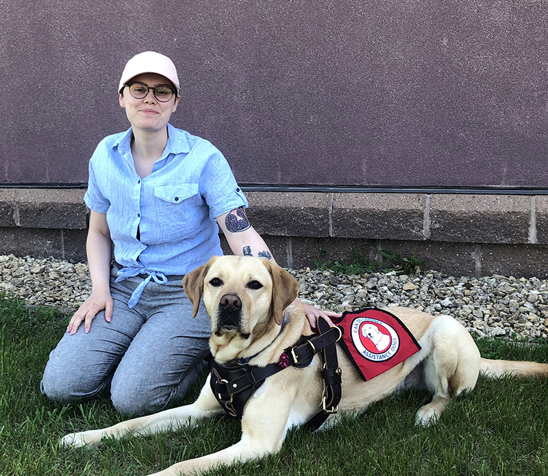 woman kneeling outside with hand on yellow Lab service dog lying next to her