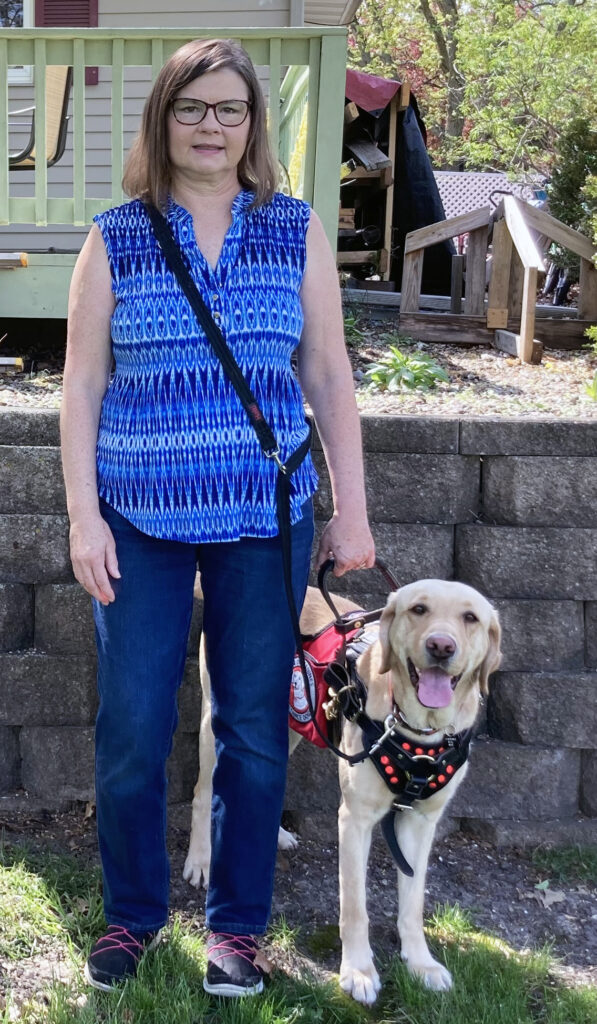 woman in blue blouse standing in front yard holding harness of service dog standing next to her