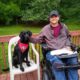 man sitting on deck in wheelchair next to black service dog on chair