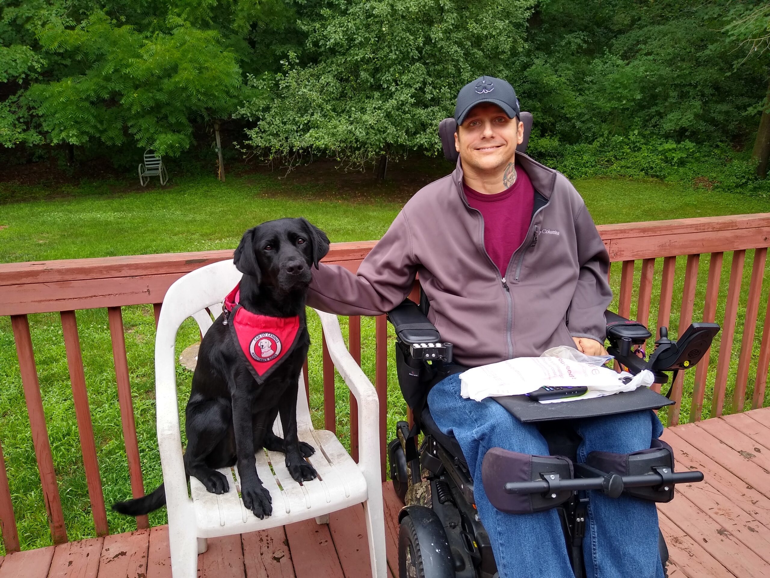 man sitting on deck in wheelchair next to black service dog on chair