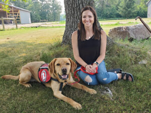 yellow Lab service dog and young woman sitting on grass in front of tree
