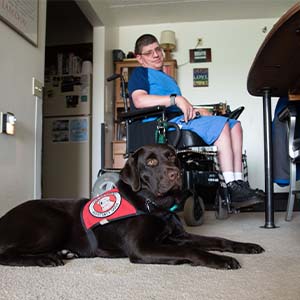 man using a motorized wheelchair is in the background of the photo with a chocolate lab wearing a red can do canines cape.