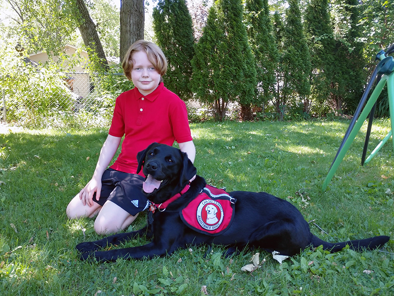 boy in red shirt sitting on grass next to black dog with service vest on