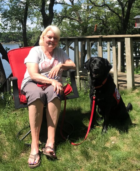 woman sitting in red chair near lake with large black dog sitting in grass beside her
