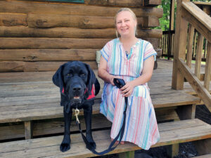 woman in striped dress sitting on log home front steps with black Lab service dog sitting beside her