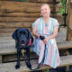 woman in striped dress sitting on log home front steps with black Lab service dog sitting beside her