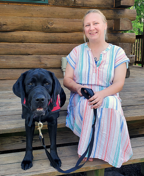 woman in striped dress sitting on log home front steps with black Lab service dog sitting beside her