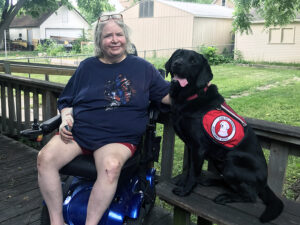 woman sitting in wheelchair on deck next to black service dog
