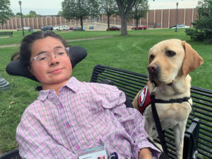 young woman sitting in powerchair with yellow Lab service dog sitting on bench next to her