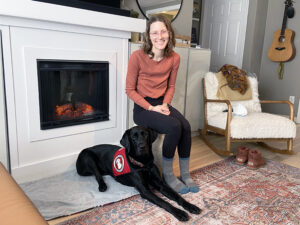 young woman sitting on chair in front of fireplace with black Lab service dog lying next to her