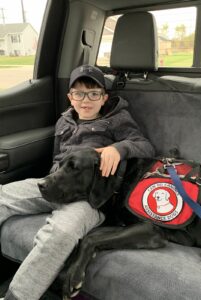 boy sitting in back seat of car, with black service dog lying its head on his lap