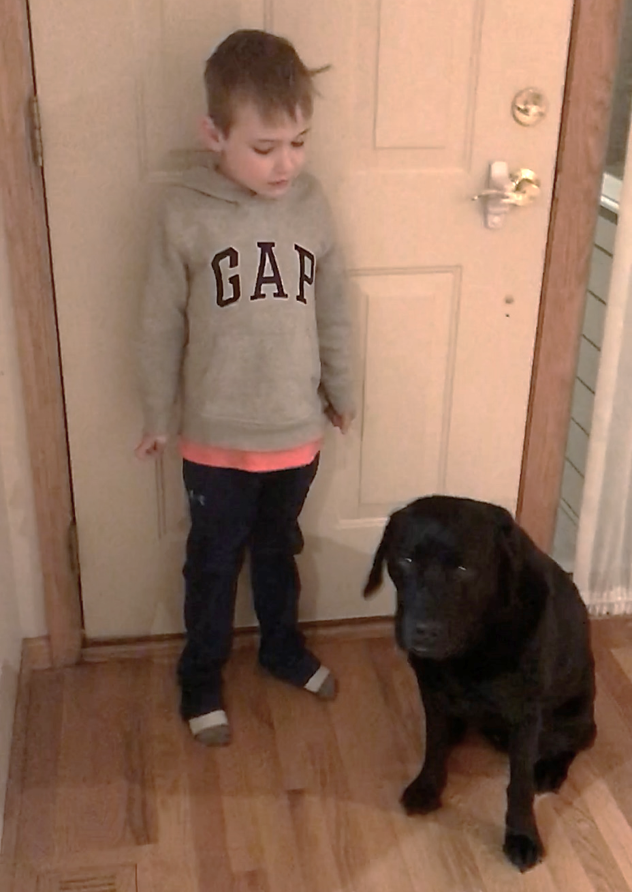 young boy standing inside front door, looking down at black Lab sitting next to him
