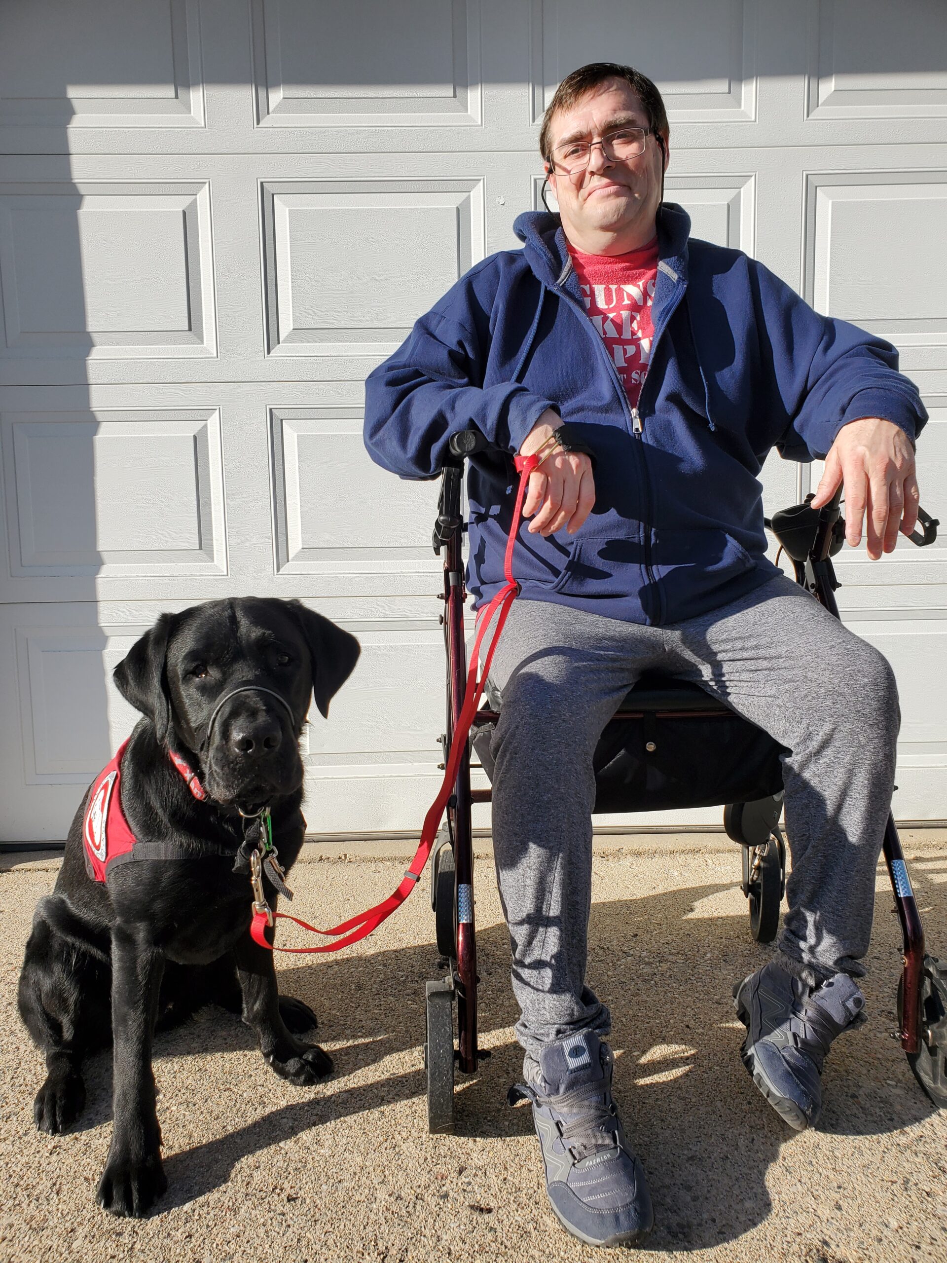 man sitting in wheelchair with black Lab Can Do Canines service dog next to him on driveway