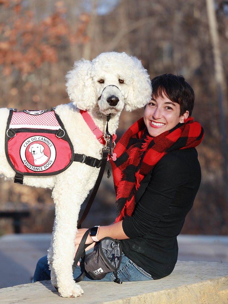 young woman sitting on concrete barrier with white poodle service dog standing next to her