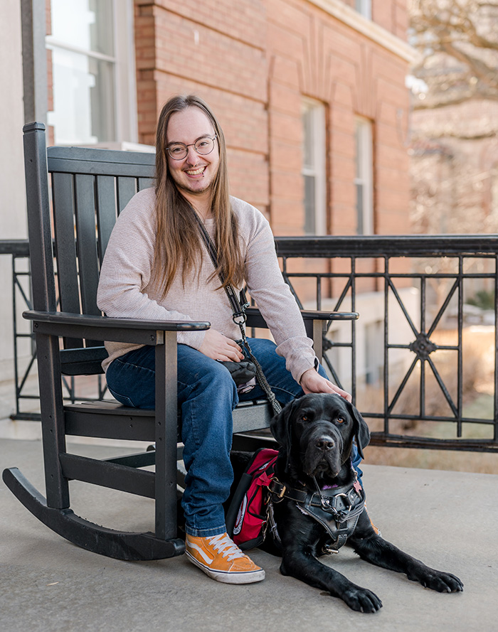 man sitting in rocking chair on building patio with black service dog lying between feet