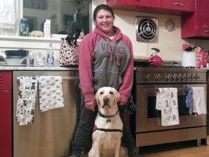 Young woman standing in kitchen with yellow Lab service dog in sitting in front of her