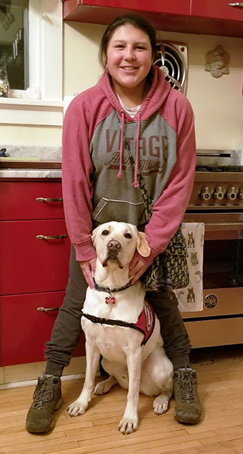 Young woman standing in kitchen with yellow Lab service dog in sitting in front of her