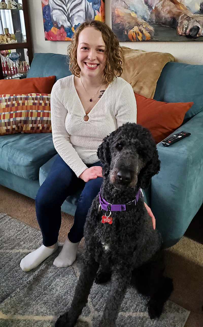 young woman sitting on edge of couch with black standard poodle service dog sitting on floor next to her