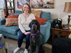 young woman sitting on edge of couch with black standard poodle service dog sitting on floor next to her