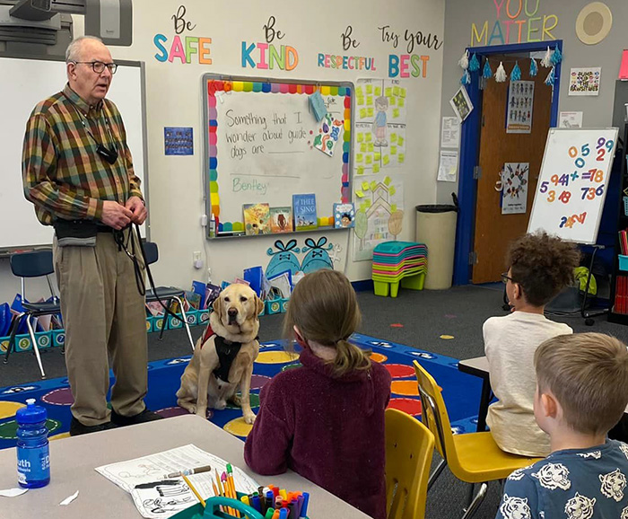 “To teach is to touch a life forever.” Most teachers are familiar with this quote by Jerry Whittle, and touching lives through teaching is something Tim seems to do without even trying. In his 53rd year as an anthropology professor at the University of Minnesota-Duluth, Tim says of teaching, “That’s been my whole life.” Something that has occurred more recently in his life–approximately 15 years ago, is a loss of hearing. That’s why when Tim’s young granddaughter insisted he needed a service dog to help him, he looked into the concept. “It’s really difficult to argue with a 7-year-old granddaughter who wants to save your life,” admits Tim. So Tim and his wife, Kim, welcomed a yellow Labrador Retriever puppy into their lives and named him Bentley. As Bentley was growing into his paws and passing obedience courses, Tim and Kim learned about Can Do Canines. Tim began the process of certifying Bentley as an owner-provided Hearing Assist Dog through Can Do Canines, and in 2018, this hearing assist team proved that what they had been taught was indeed certification-worthy. Bentley was trained to alert Tim to a wide variety of sounds, and Tim figures that his loyal Lab has saved his life, just by literally touching him at least 15 times so far. He describes the first time that Bentley jumped into major alert action was when Tim was sharing his story at a Can Do Canines Board of Directors meeting. A fire alarm went off, and Bentley was adamant that Tim needed to make his way to an exit. Remembering that it was a cold winter day, and the group was fairly certain that it was a false alarm, Tim recalls, “I was hoping we would get to that first door, and [Bentley] would let me stay inside. But no, he headed right through, down the sidewalk and across the parking lot to the far side to safety.” Bentley also alerts to more common sounds like oven timers, doorbells, door knocks, car doors closing and unusual human sounds in the yard. The feeling of security Bentley brings extends beyond Tim’s own comfort. He explains, “The peace of mind that Bentley brings to my wife and to the whole family is extremely important.” Kim travels frequently, and Tim confesses, “I sleep like a log. . .She can now go away and is fully confident that if some emergency comes up, Bentley will take care of it.” In fact, Tim stresses Bentley’s persistence, saying “He doesn’t have a snooze alarm. He gets me up…and he’s all business.” Since Tim can only wear his cochlear implants about four to five hours per day, it’s a good thing Bentley's work ethic is so strong. In any potentially dangerous circumstance, Bentley always reacts with Tim’s best interest in mind. “His concern is not to run away, but to take me away from that situation,” says Tim. Some of the times when Bentley has had to alert Tim is when Tim is in teaching mode, and Bentley has logged many days at the university campus over the years. However, given Tim’s passion to educate others, he has also made it a practice to share with others the benefits of aa assistance dog. In April, the pair presented to a first-grade class at Lowell Elementary School in Duluth. Tim says, “There’s nothing like visiting and teaching first graders. They’re just so excited. When you ask if there’s a question, every hand shoots up.” Tim has also shared his important message at the Cloquet Library and plans to give a presentation at the Duluth Children’s Museum soon. Whenever and wherever Tim is educating others, Bentley is a necessary part of his life. He says, “When I’m teaching, now I can be fully independent.” And with that teaching, whether it’s about anthropology or assistance dogs, more lives will be touched.