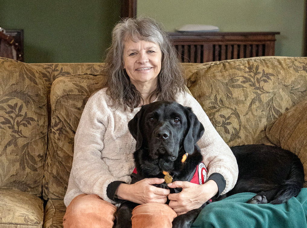 woman sitting on sofa, with black Lab service dog lying on her leg