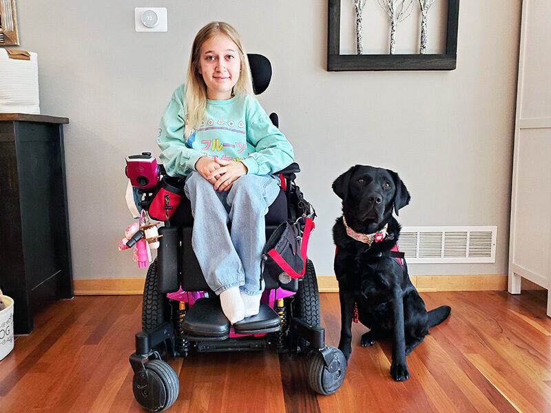 young woman sitting in wheelchair with black Lab sitting on floor beside her