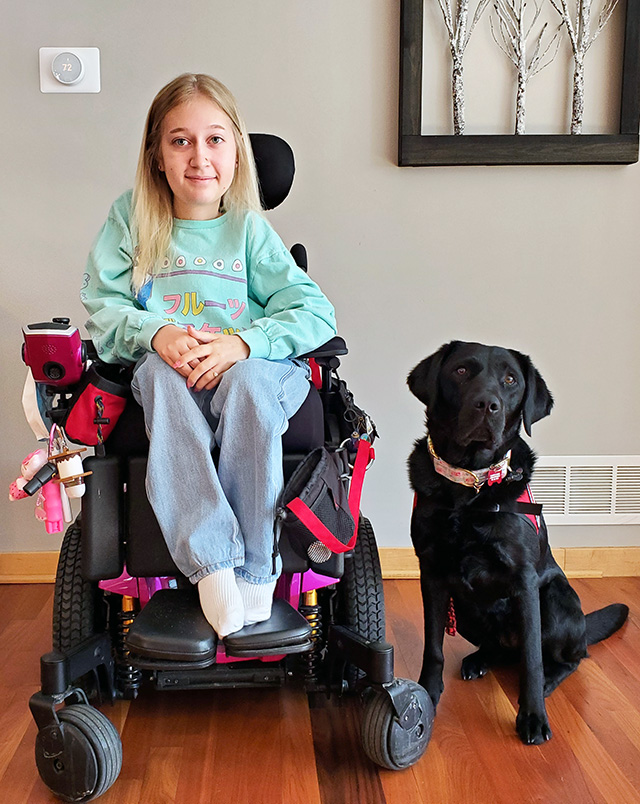 young woman sitting in wheelchair with black Lab sitting on floor beside her