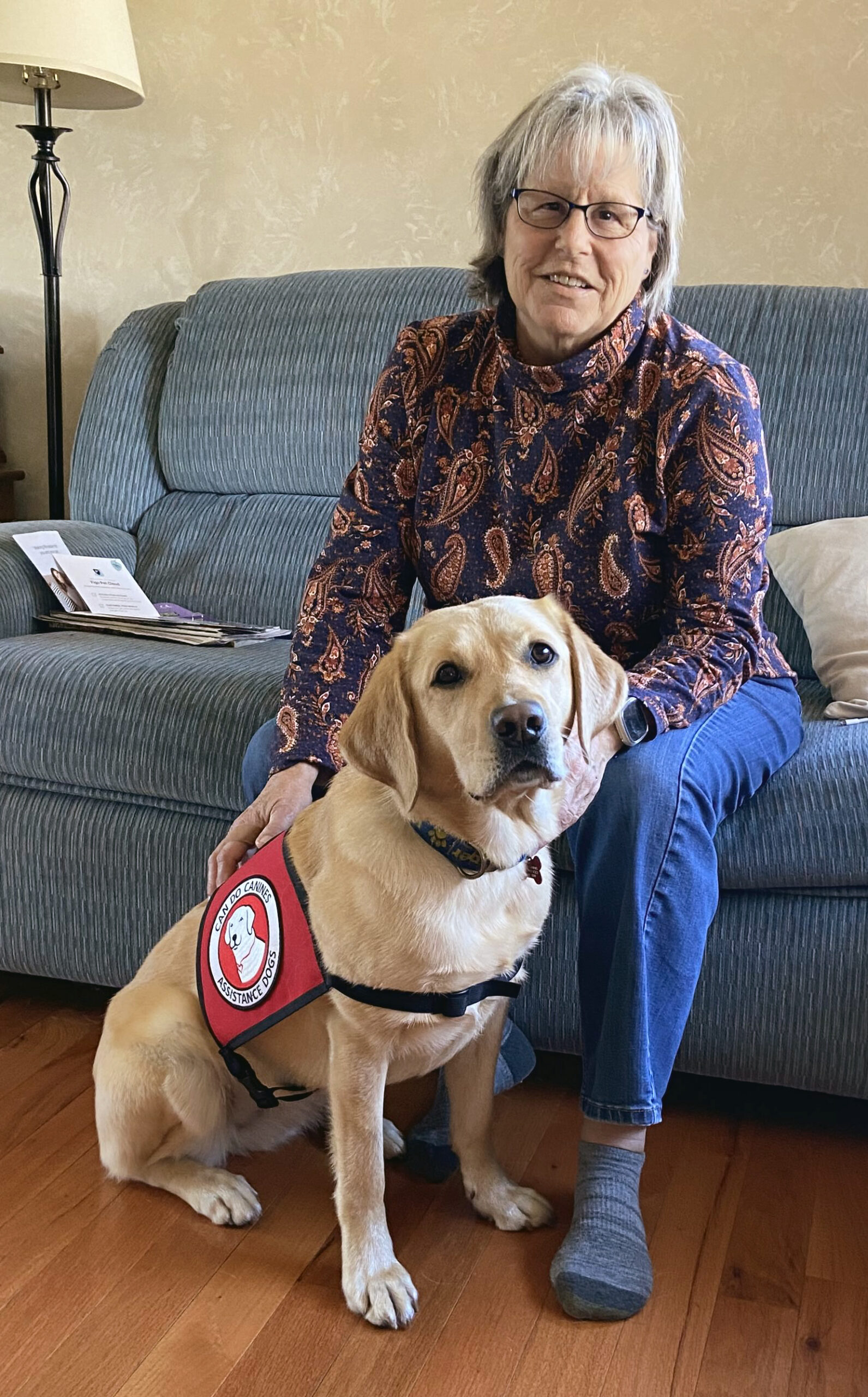 woman sitting on couch with service dog wearing Can Do Canines cape sitting in front of her