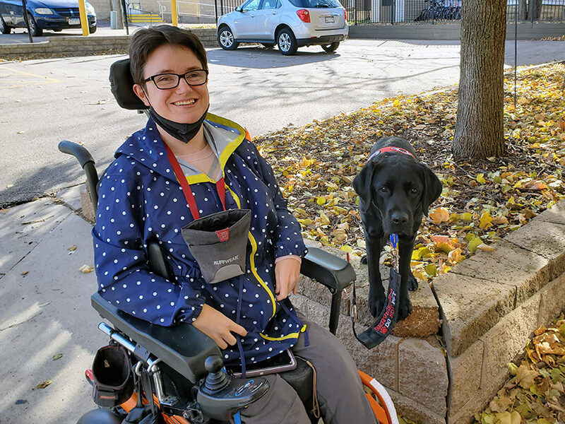 young woman sitting in powerchair on sidewalk with black Lab service dog sitting on high curb next to her