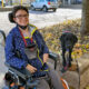 young woman sitting in powerchair on sidewalk with black Lab service dog sitting on high curb next to her