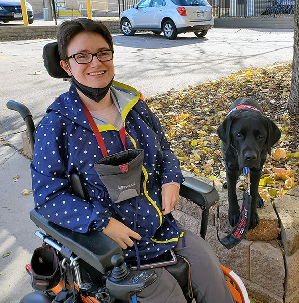 young woman sitting in powerchair on sidewalk with black Lab service dog sitting on high curb next to her