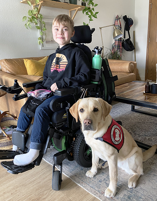 blond boy sitting in wheelchair with yellow Lab service dog sitting next to him