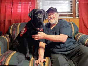 man with beard and glasses sitting on couch with black Lab service dog sitting up next to him