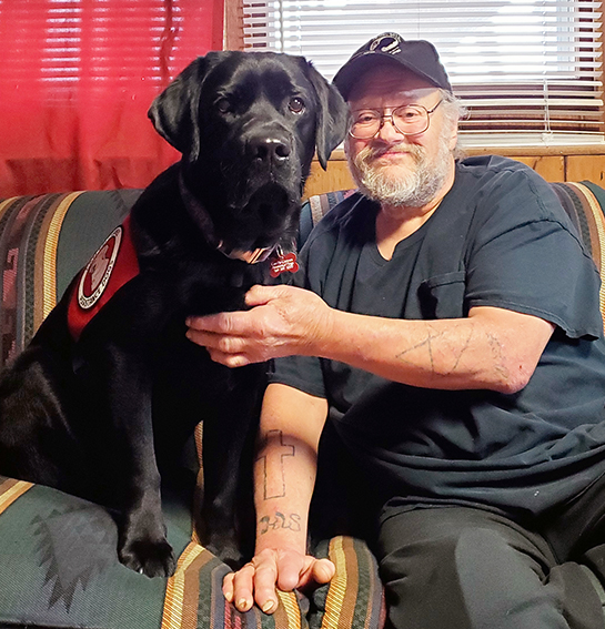 man with beard and glasses sitting on couch with black Lab service dog sitting up next to him