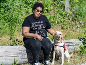 woman sitting on fallen log with yellow Lab service dog sitting next to her