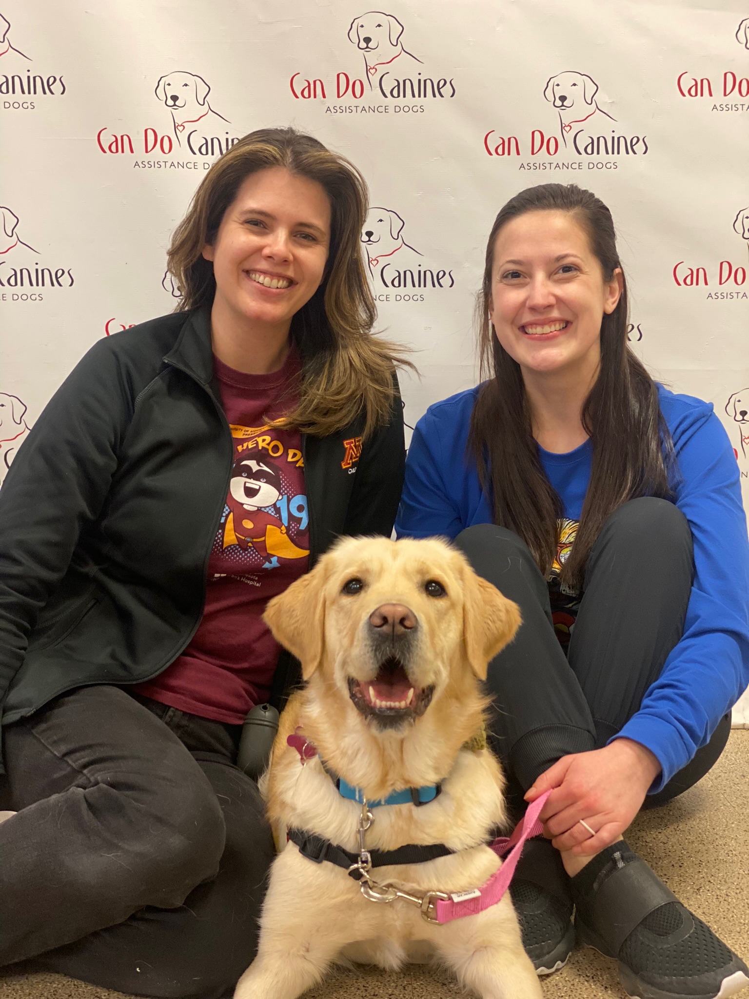 two woman sitting on each side of yellow Lab service dog in front of backdrop for Can Do Canines