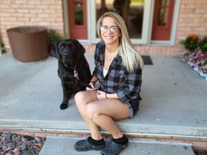 young woman with long blond hair sitting on front step with black Lab
